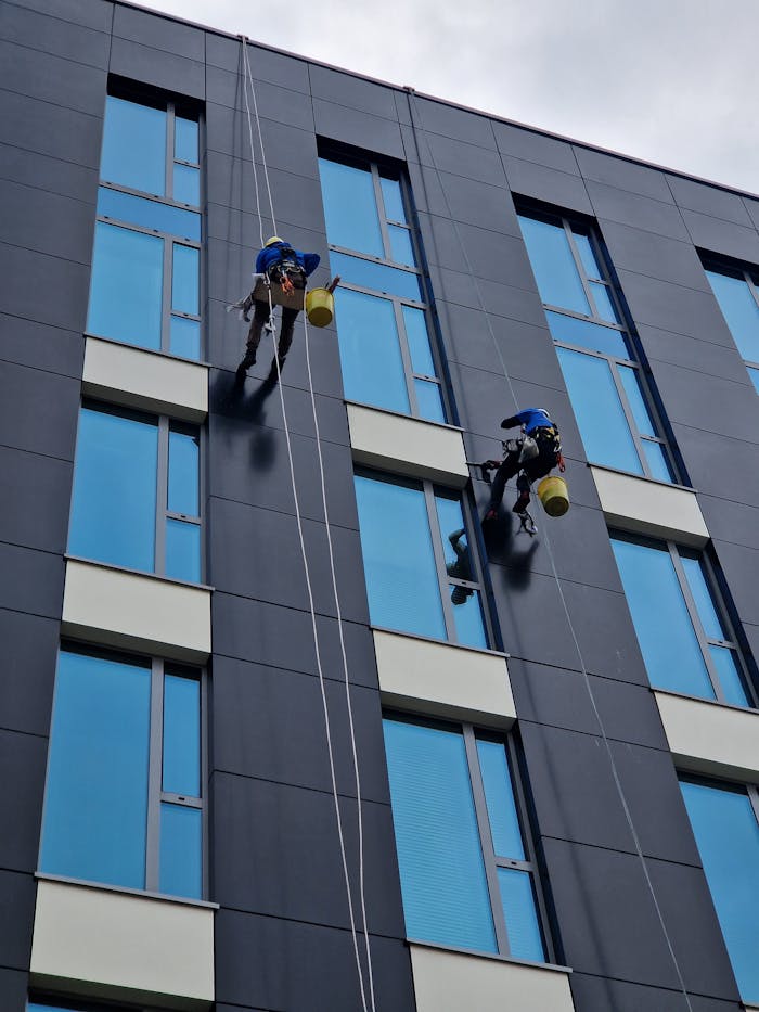 Two workers clean a modern building facade at height with safety ropes.