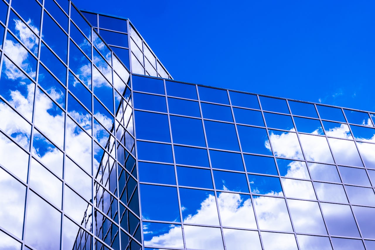 Low angle view of a modern glass building reflecting clouds against a vivid blue sky.